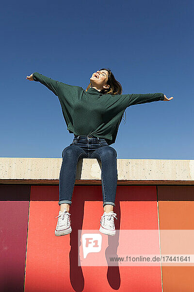 Carefree woman with arms outstretched sitting on retaining wall during sunny day