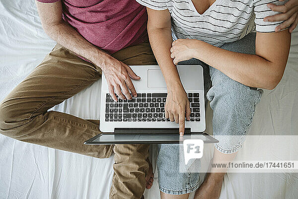 Woman touching screen while sitting with man using laptop on bed
