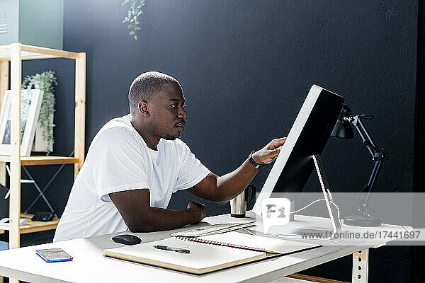 Male freelance worker touching desktop screen while sitting at desk in studio