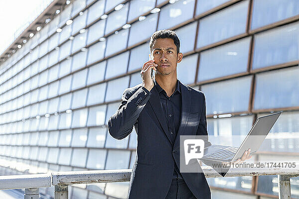 Young male professional with laptop talking on mobile phone while standing by railing