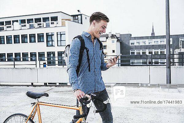 Cheerful man with bicycle using mobile phone while walking on parking garage rooftop