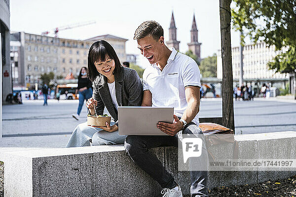 Happy male and female friends using laptop while sitting on retaining wall