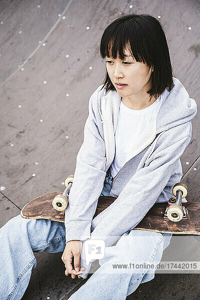 Female teenager with skateboard sitting on sports ramp
