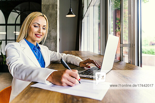 Lächelnde Geschäftsfrau schreibt auf Papier  während sie mit Laptop am Schreibtisch im Büro sitzt