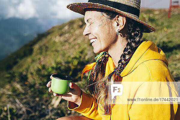 Fröhliche Frau mittleren Alters mit Hut sitzt an einem sonnigen Tag auf einem Berg