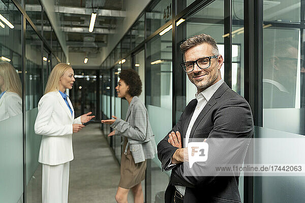Smiling businessman standing with arms crossed while female colleagues discussing in corridor at office