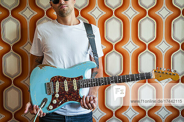 Young male musician holding guitar in front of wall