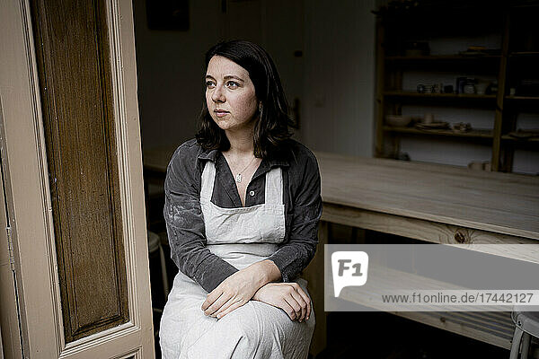 Thoughtful craftswoman wearing apron sitting in workshop