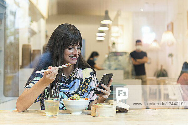 Smiling mid adult woman using smart phone while eating poke in restaurant