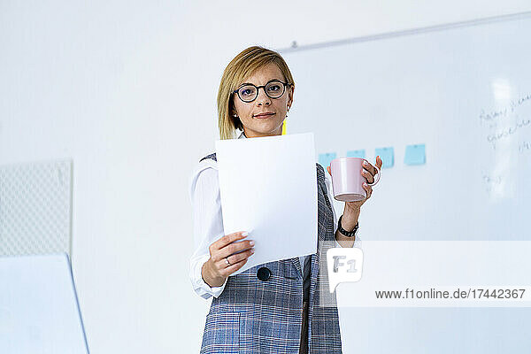 Businesswoman with document and coffee mug standing by whiteboard in office