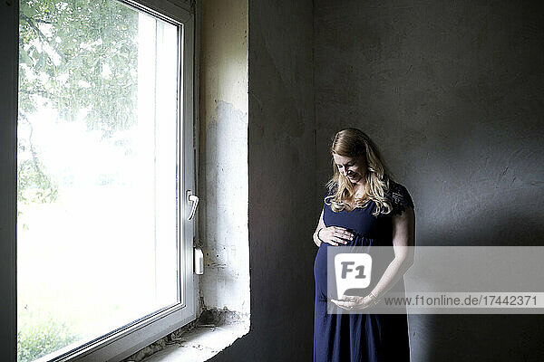 Pregnant woman touching abdomen while standing by window