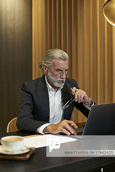 Mature businessman holding eyeglasses while working on laptop in hotel