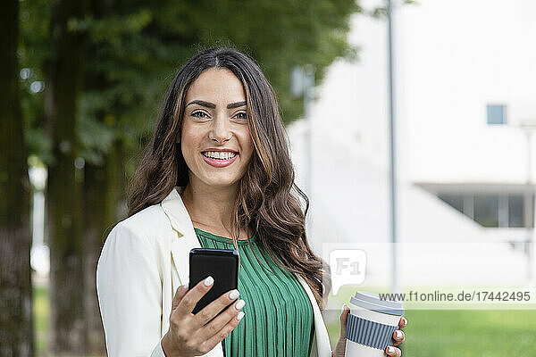 Happy young woman holding mobile phone and disposable coffee cup