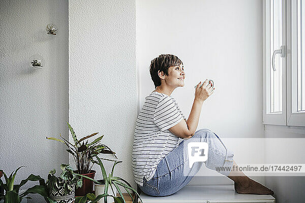 Smiling mid adult woman holding mug while sitting by plants at home