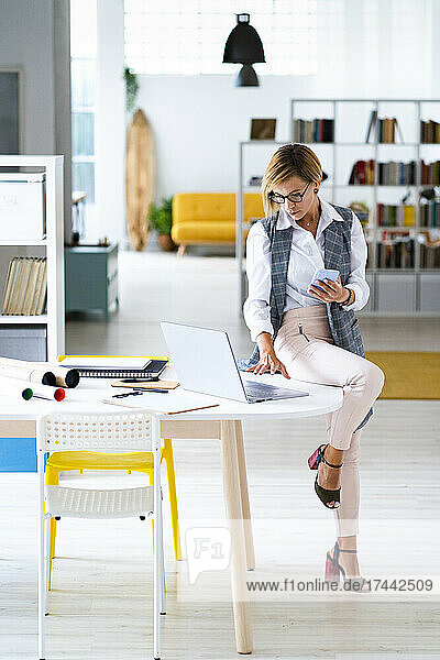 Businesswoman with smart phone using laptop while sitting on desk