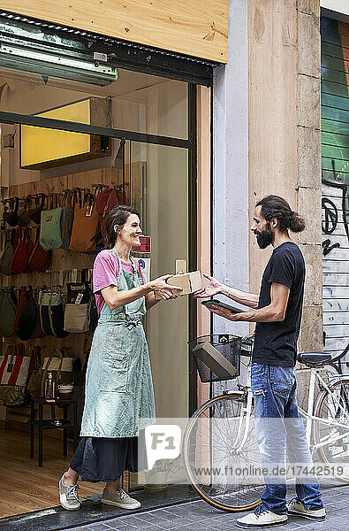 Smiling female entrepreneur giving package to male delivery person while standing outside store