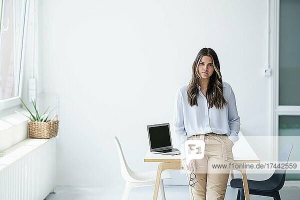 Young businesswoman with hand in pocket leaning on desk