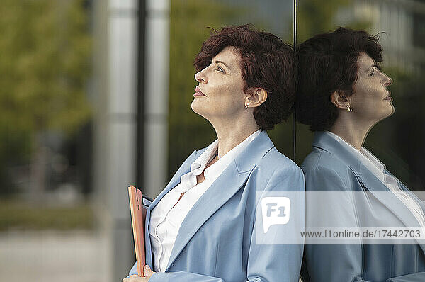 Thoughtful businesswoman holding file while leaning on glass wall