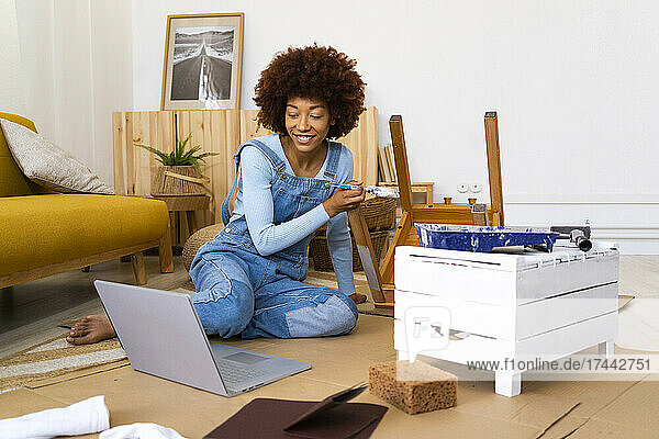 Afro woman using laptop while painting chair in living room