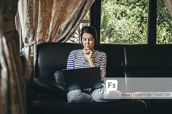 Woman with hand on chin using laptop on couch at home