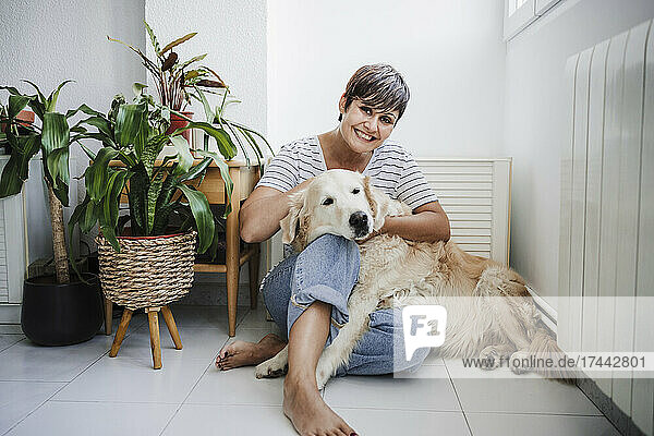 Lächelnde Frau sitzt mit Hund zu Hause neben Pflanzen