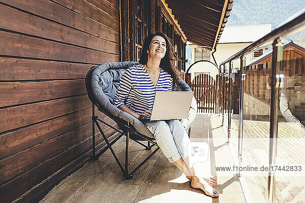 Lächelnde Frau schaut nach oben  während sie auf einem Stuhl mit Laptop auf dem Balkon sitzt