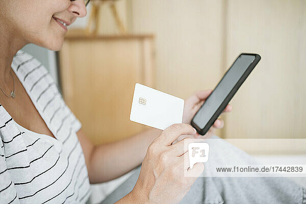 Frau hält Kreditkarte in der Hand  während sie zu Hause ihr Smartphone benutzt