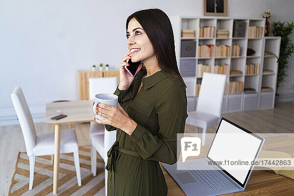 Young businesswoman having coffee while talking on smart phone at home