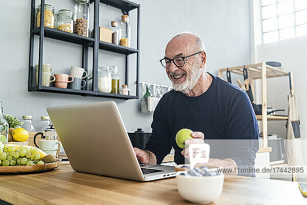 Glücklicher älterer Mann hält Apfel in der Hand  während er zu Hause Laptop benutzt