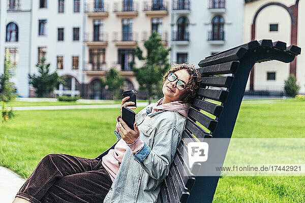 Mid adult woman with mobile phone having coffee while sitting on bench