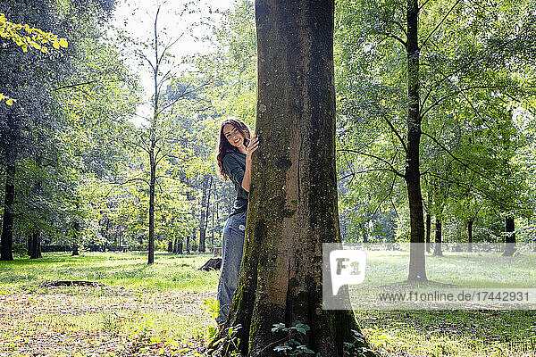 Smiling woman standing behind tree trunk in park