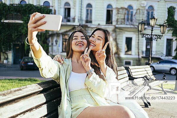 Female friends gesturing peace sign while taking selfie through smart phone in city