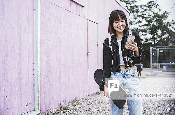 Smiling teenage girl with skateboard using smart phone on footpath