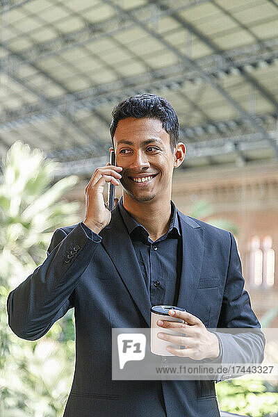 Happy young businessman holding disposable coffee cup talking on mobile phone