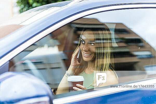 Smiling woman with coffee cup talking on smart phone in car
