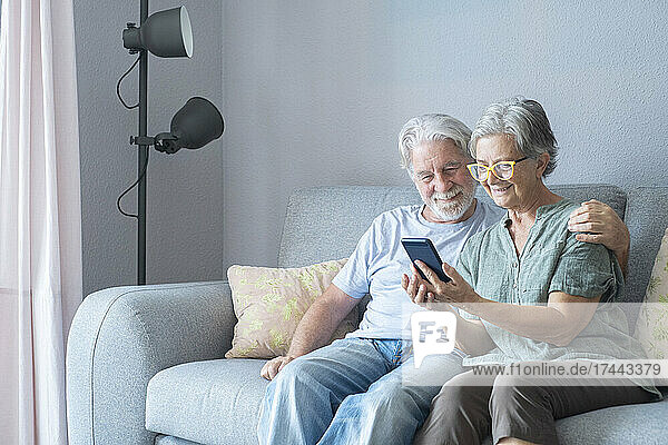 Lächelnde ältere Frau  die ihr Mobiltelefon mit einem Mann auf dem Sofa teilt