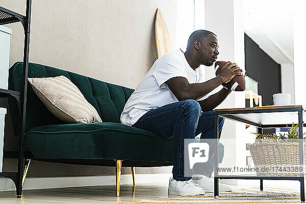 Nachdenklicher Mann mittleren Alters mit Rugbyball sitzt zu Hause auf dem Sofa