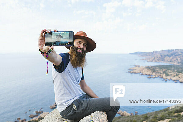 Bearded man taking selfie through smart phone while sitting on rock