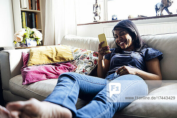 Lächelnde Frau surft per Smartphone im Internet  während sie zu Hause auf dem Sofa sitzt