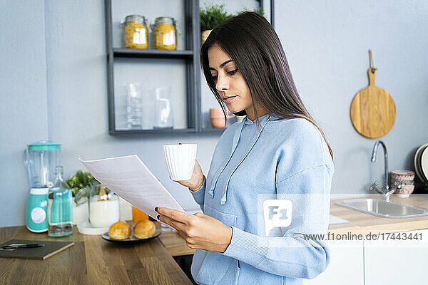 Beautiful young woman reading document while having coffee at home