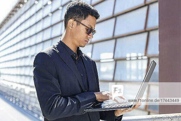 Male professional typing on laptop while standing outside office building