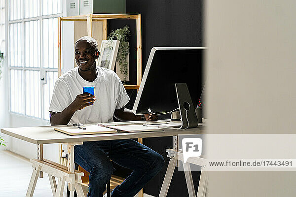 Smiling male freelancer holding mobile phone while sitting at desk in studio