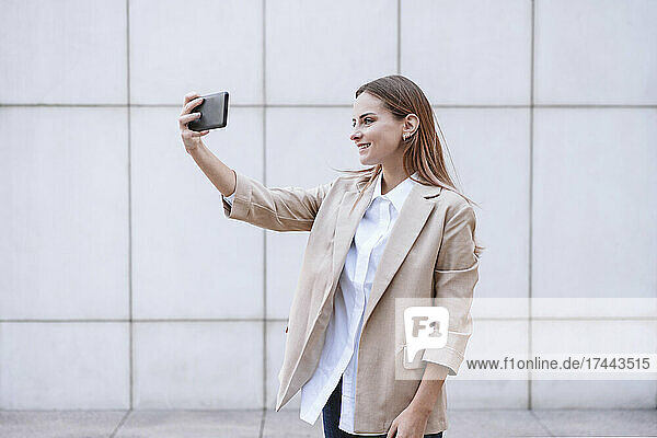 Young businesswoman taking selfie through smart phone in front of wall