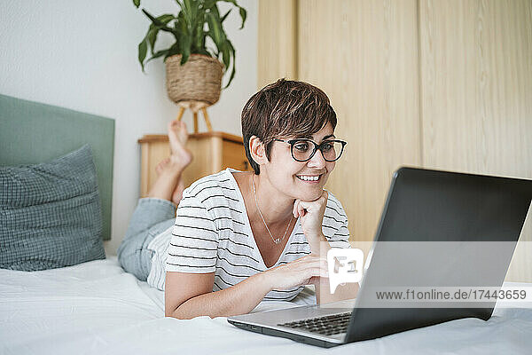 Woman with hand on chin using laptop while lying on bed at home