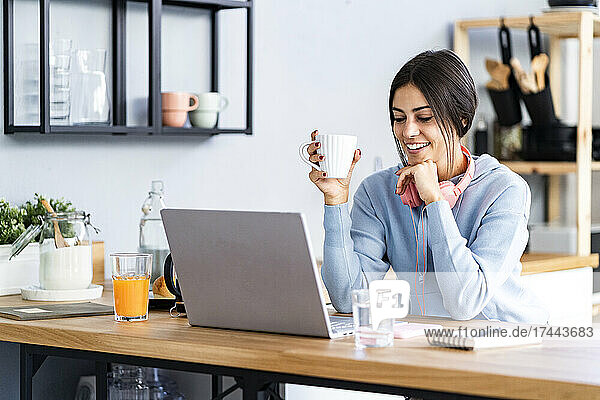 Smiling beautiful woman with hand on chin looking at laptop while sitting in kitchen