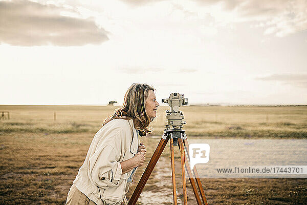 Mature woman looking through theodolite at field