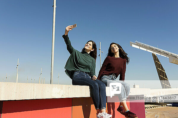 Frau macht Selfie  während sie mit Freundin auf Stützmauer sitzt