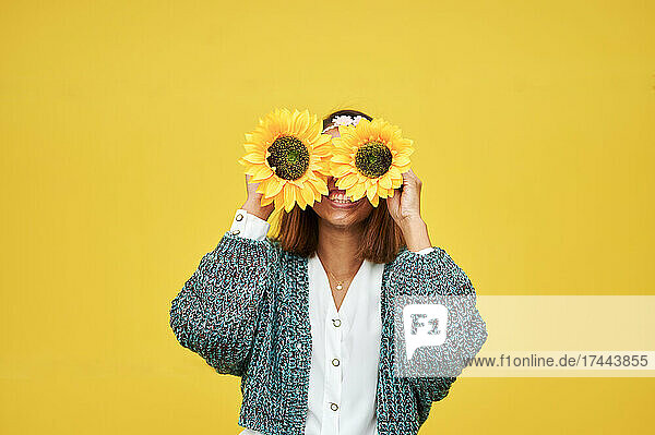 Young woman covering face with sunflowers in front of wall