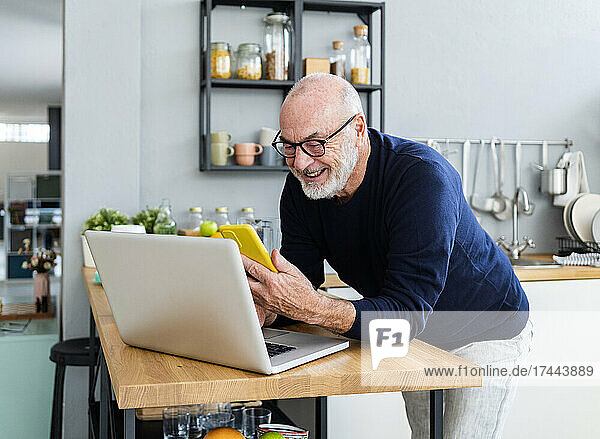 Smiling man with laptop using mobile phone in kitchen