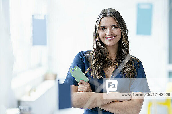 Smiling female business professional standing with arm crossed seen through glass wall